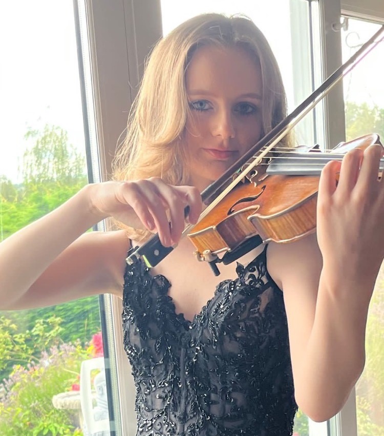 Abigail is a teenager. She has long hair and is wearing a black dress. She is posed playing a violin as she looks into the camera. Behind her, windows overlook a lawn and trees.