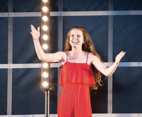 Grace is a teeanger. She is wearing a red strappy top and flowing red trousers as she stands on a lit stage, arms outspread, singing.