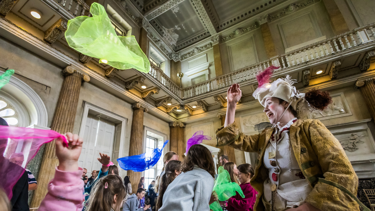 People toss colorful scarves in a sunlit room inside Wentworth Woodhouse, surrounded by grand columns and ornate ceilings.