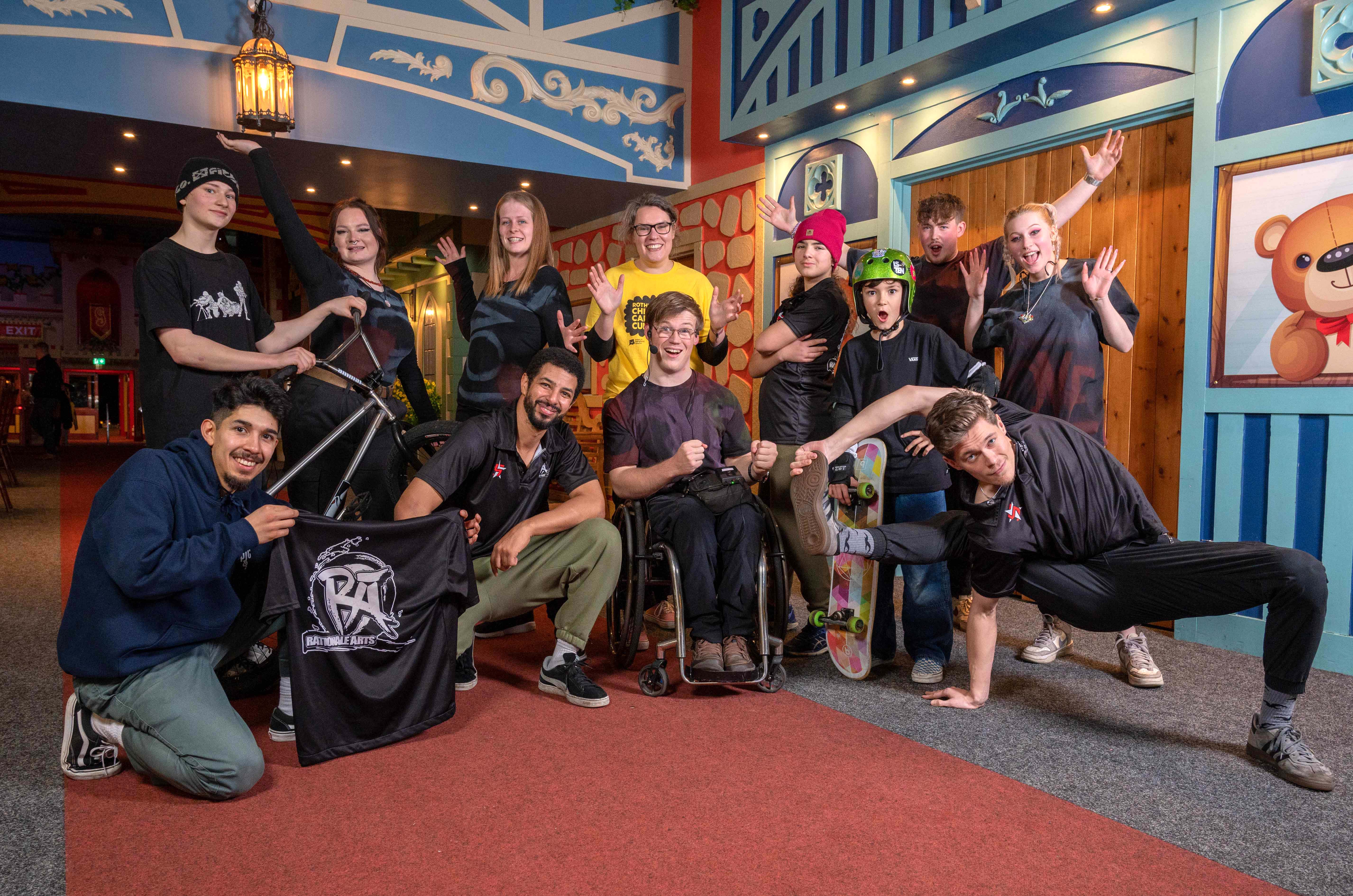 A group of twelve smiling people pose for the camera. Some of them are dressed in skater clothes, and wearing helmets and holding bikes or skateboards. Some are in break dancing poses.