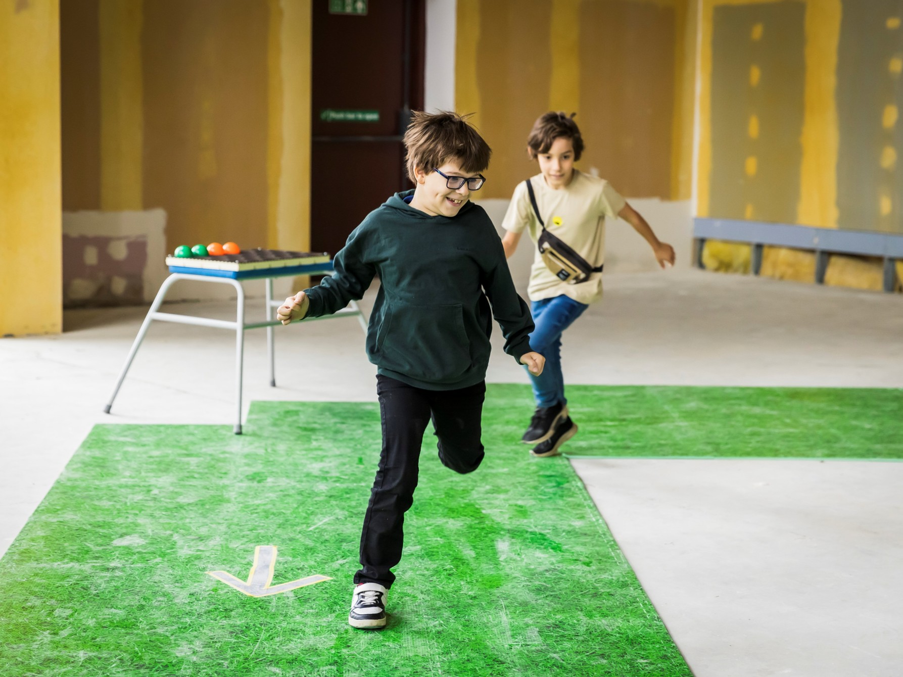 Two children run on green matting