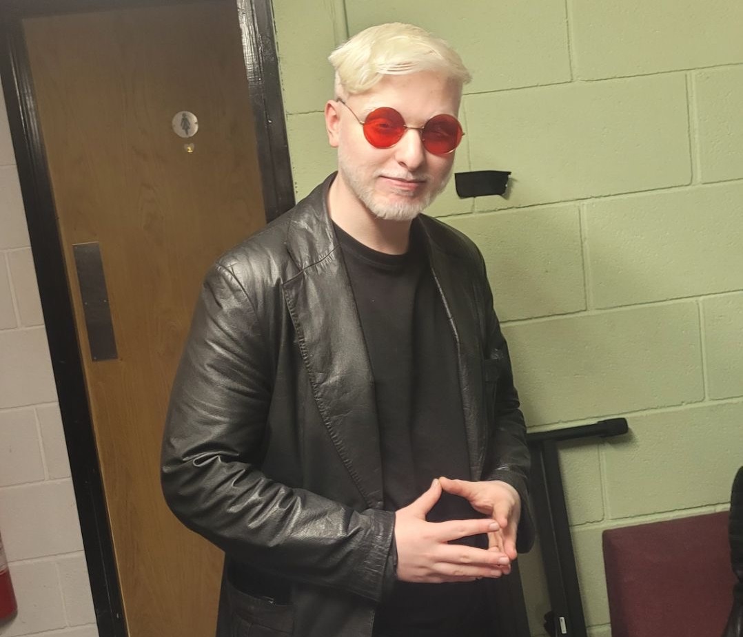 Steve is a teenager. He looks at the camera with a slight smile. He is wearing red tinted sunglasses, a black leather jacket, and a black tee-shirt.